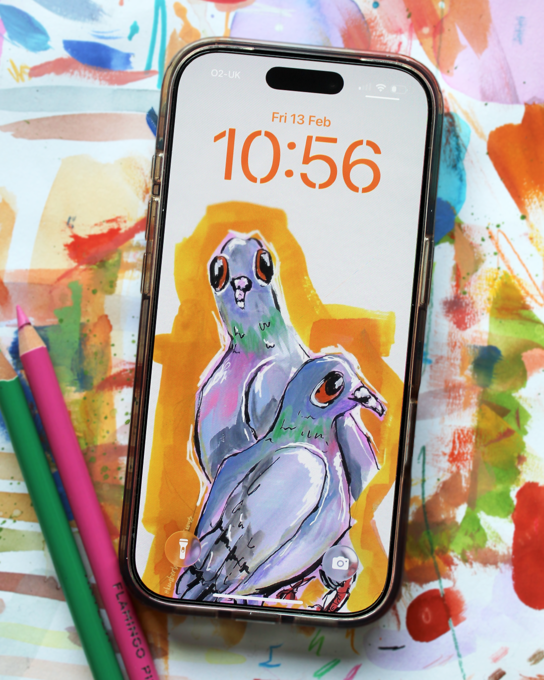iPhone with a colorful screen showing two pigeons, surrounded by art supplies on a multicolored background
